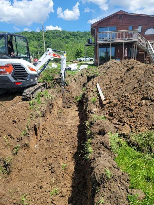 Compact excavator cutting a long, deep trench down a slope beside a two-story house