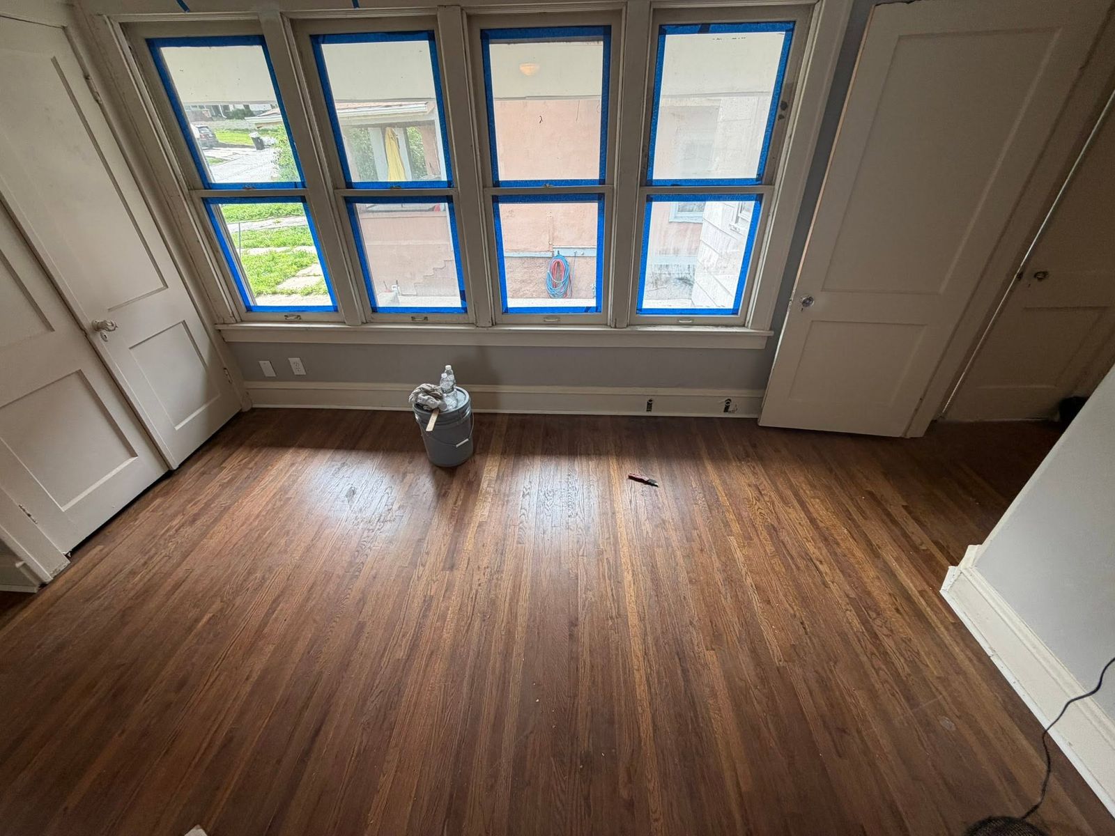 Hardwood floor mid-refinish, windows taped off, in a bright room with a bank of windows