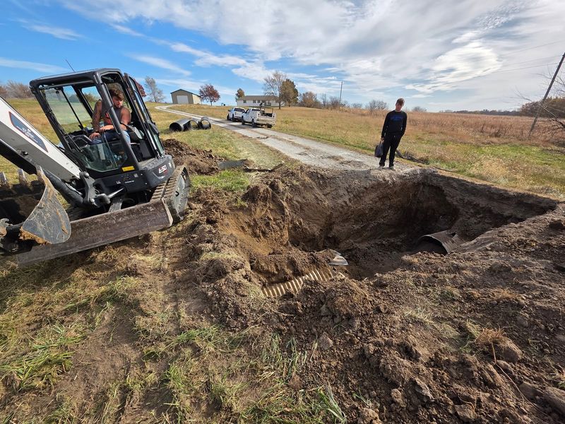 Compact track loader digging out a hole for a culvert, corrugated pipe staged nearby