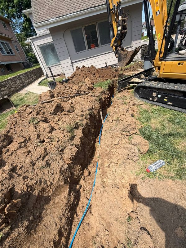 Compact excavator trenching across a front yard with blue conduit in the trench