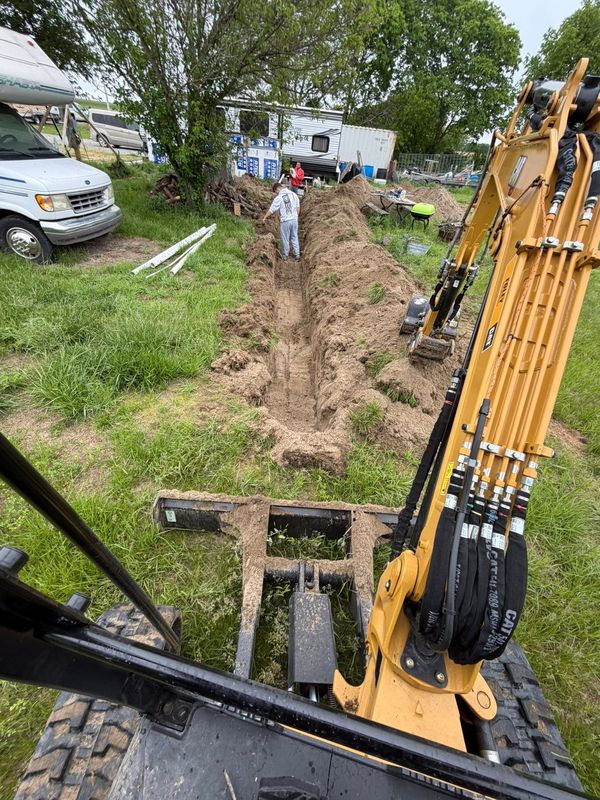 Excavator cab view trenching across a grassy rural lot